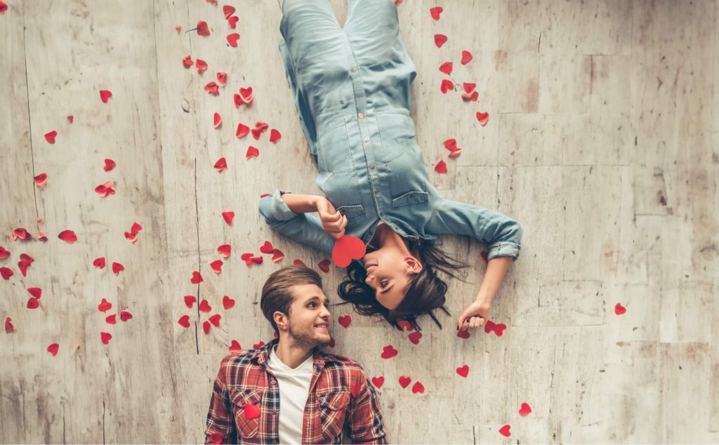 Couple allongé au sol entouré de pétales de cœur rouge, la femme tenant un cœur en papier rouge, regard amoureux échangé, ambiance romantique et tendre sur fond de parquet clair qui ressemble à du parquet. La femme à une combinaison en jeans et l'homme porte une chemise à carreaux rouge