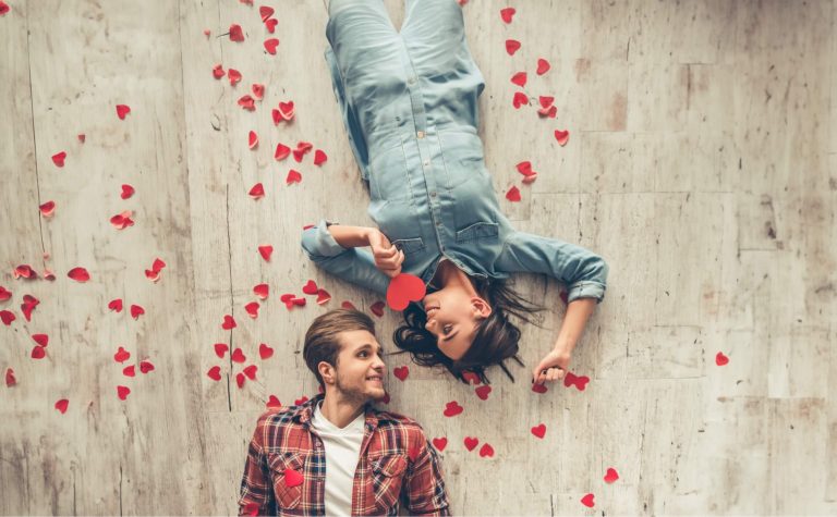 Couple allongé au sol entouré de pétales de cœur rouge, la femme tenant un cœur en papier rouge, regard amoureux échangé, ambiance romantique et tendre sur fond de parquet clair qui ressemble à du parquet. La femme à une combinaison en jeans et l'homme porte une chemise à carreaux rouge