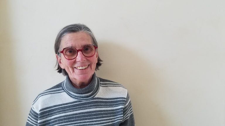 Portrait d’une femme souriante aux cheveux gris courts, portant des lunettes rouges rondes et un pull à col roulé rayé, posant devant un mur clair