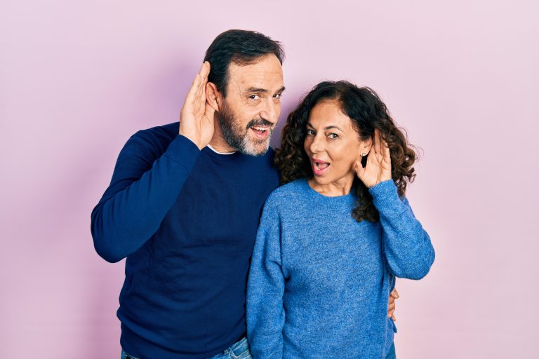 Couple mature souriant et faisant semblant d’écouter en tenant une main près de l’oreille, posant avec humour devant un fond rose, habillés en bleu