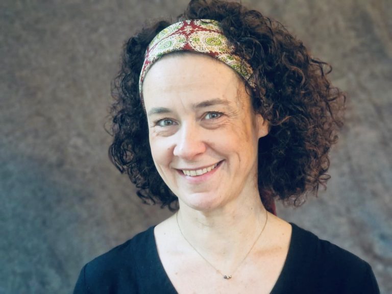 Portrait d’une femme souriante aux cheveux bouclés, portant un bandeau coloré et un haut noir, posant devant un fond neutre