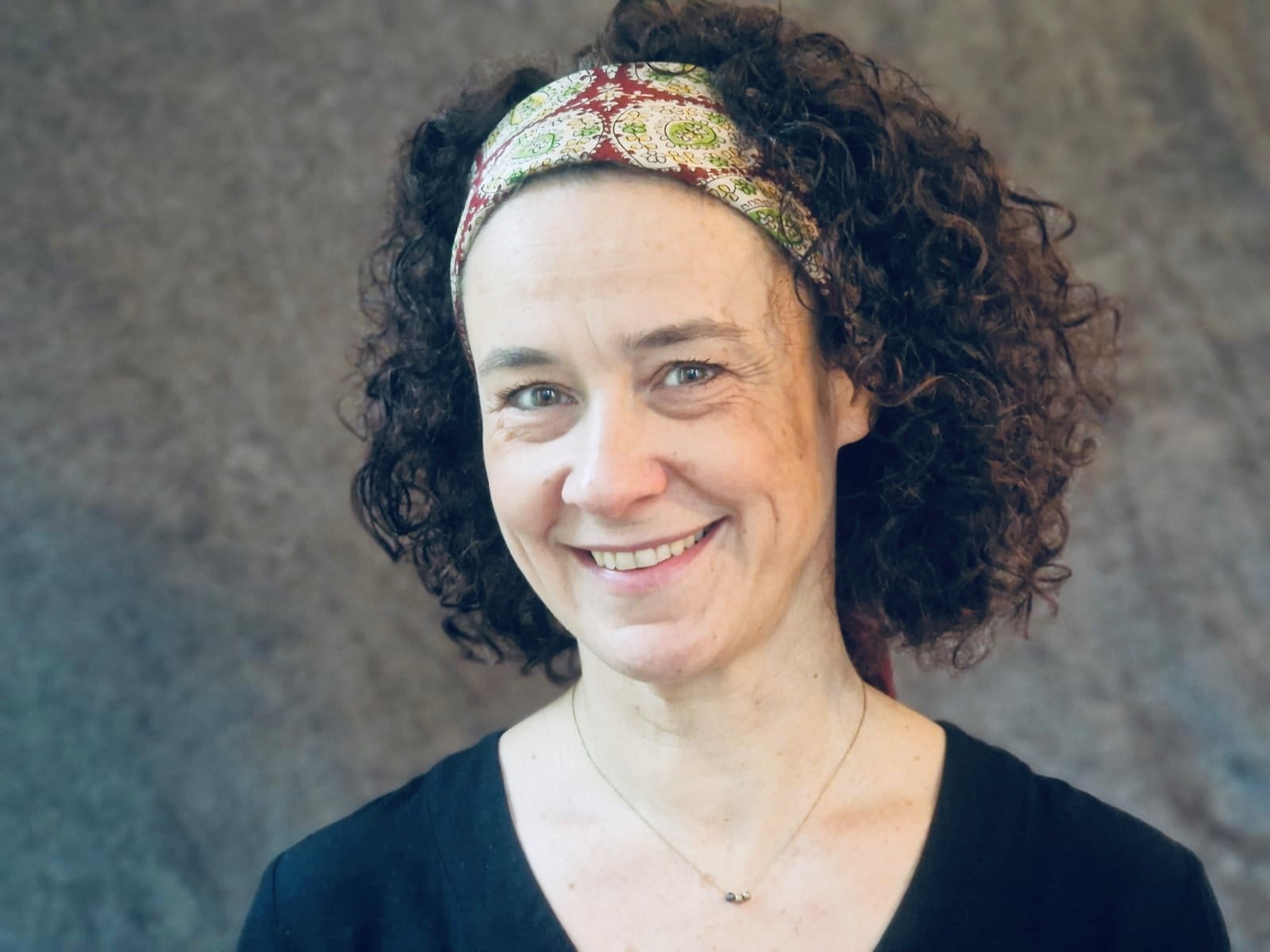 Portrait d’une femme souriante aux cheveux bouclés, portant un bandeau coloré et un haut noir, posant devant un fond neutre
