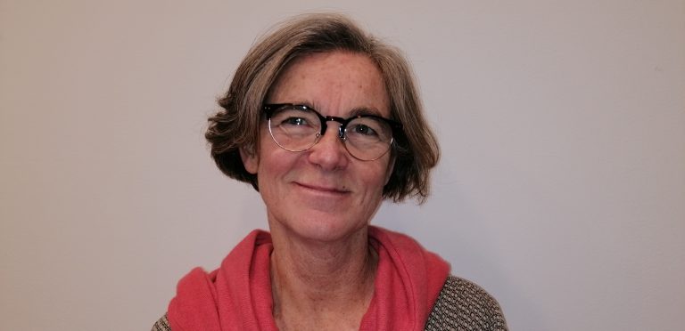 Portrait d’une femme souriante aux cheveux gris courts, portant des lunettes rondes et une veste avec un pull rose, sur fond clair