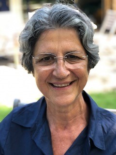 Dominique est une femme plutôt âgée, souriantes ayant des lunettes de couleur grise. Elle a des cheveux courts de couleurs sel et poivre et porte un chemisier bleu.