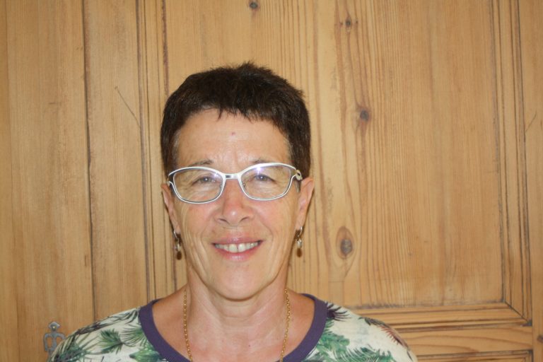 Portrait d’une femme souriante aux cheveux courts, portant des lunettes blanches et un haut à motifs végétaux, posant devant un mur en bois