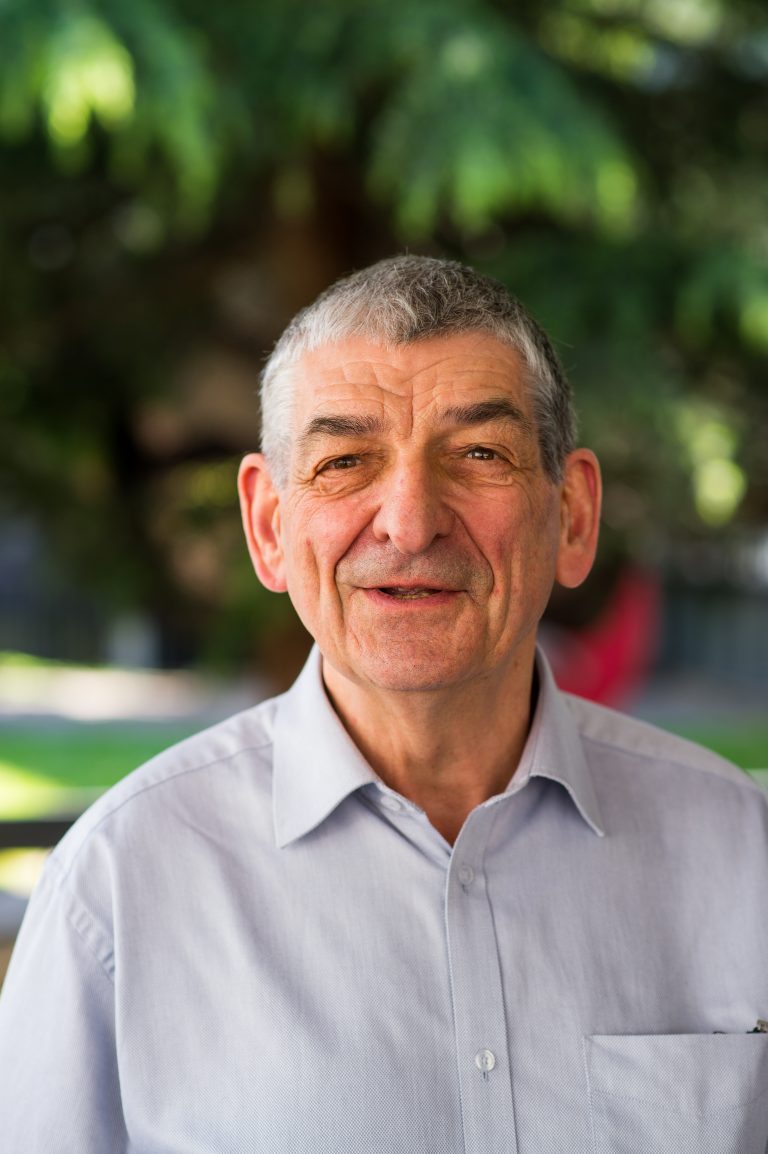 Portrait d’un homme souriant aux cheveux gris, portant une chemise claire, photographié en extérieur avec un fond d’arbres et de verdure