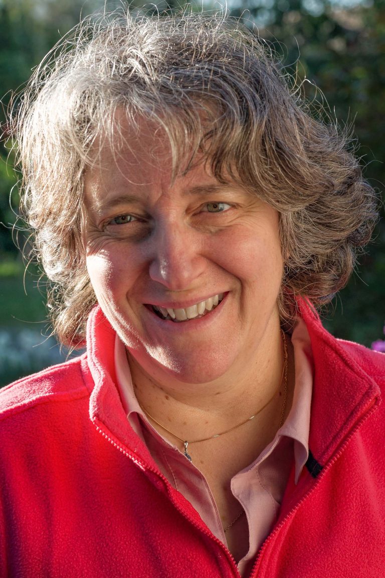 Femme souriante aux cheveux gris courts portant une veste polaire rose, en extérieur avec lumière naturelle