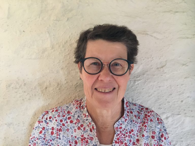 Portrait d’une femme souriante aux cheveux courts bruns, portant des lunettes rondes et une chemise fleurie, devant un mur clair en pierre