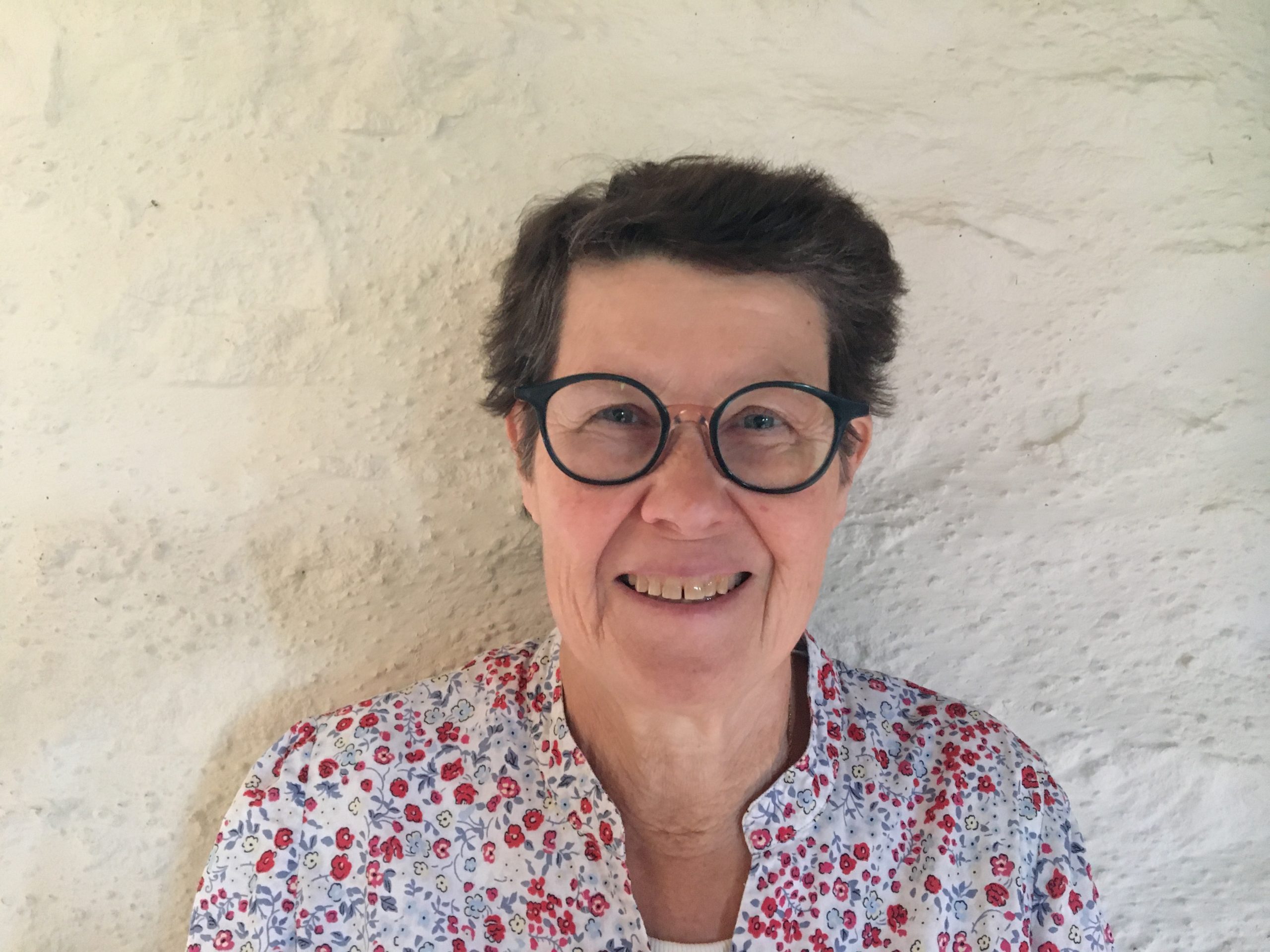 Portrait d’une femme souriante aux cheveux courts bruns, portant des lunettes rondes et une chemise fleurie, devant un mur clair en pierre