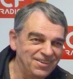 Portrait d’un homme souriant aux cheveux gris, photographié devant un logo de radio, en contexte d’interview ou d’intervention médiatique