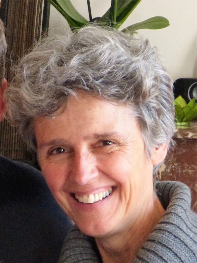 Femme souriante aux cheveux courts et gris, en intérieur, devant une plante verte