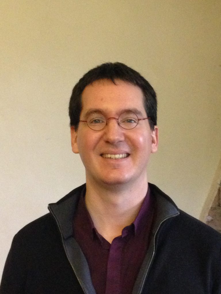 Portrait d’un homme souriant portant des lunettes rondes, une chemise violette et un gilet zippé noir, devant un mur beige