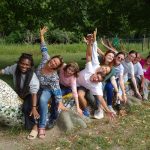 Groupe joyeux de personnes assises sur une sculpture colorée en forme de serpent, penchées vers l'avant avec les bras levés, dans un parc verdoyant