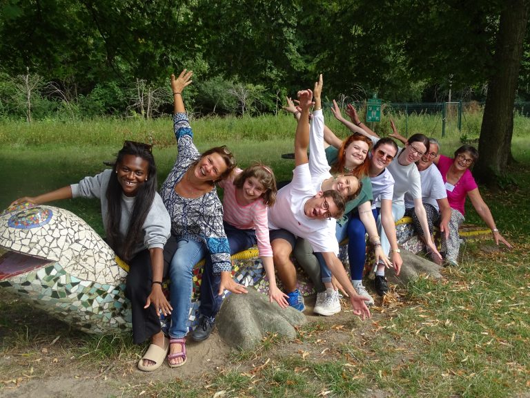 Groupe joyeux de personnes assises sur une sculpture colorée en forme de serpent, penchées vers l'avant avec les bras levés, dans un parc verdoyant