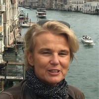 Portrait d'une femme parlant avec des lunettes de soleil et une coupe de cheveux blond et courte. Elle porte également un foulard bleu avec des points blancs. En arrière blanc il y a des bateau qui navigue sur une rivère.