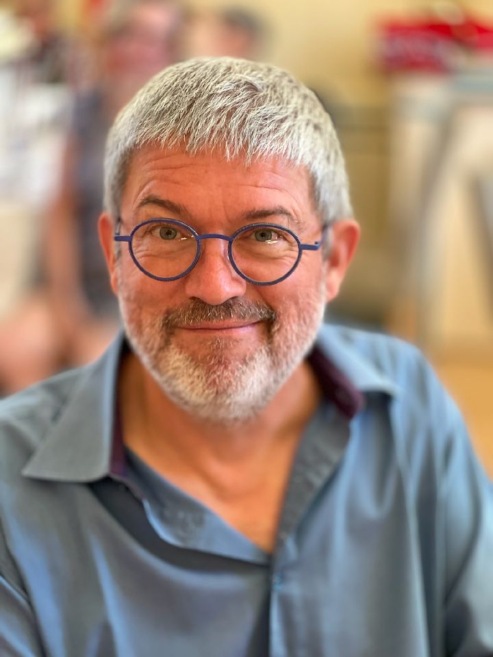 Portrait d'un homme souriant avec un air chaleureux. Il porte des lunettes de couleur bleue, une barbe et des cheveux courts de couleur grise. Il porte également une chemise bleu avec les 2 boutons du col légèrement ouvert