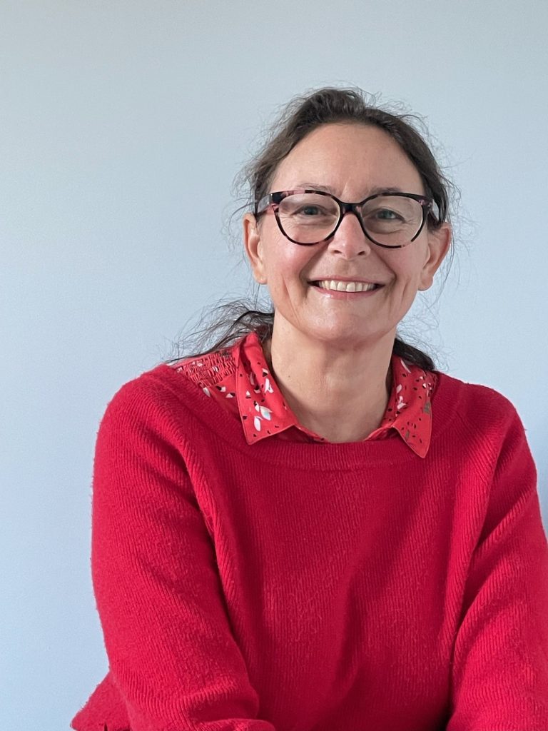 Portrait d'une femme souriante avec des cheveux bruns attachés, portant des lunettes à monture noire et un pull rouge à col rond avec des motifs décoratifs sur le col. Elle se tient devant un fond uni de couleur claire, offrant une expression chaleureuse et accueillante