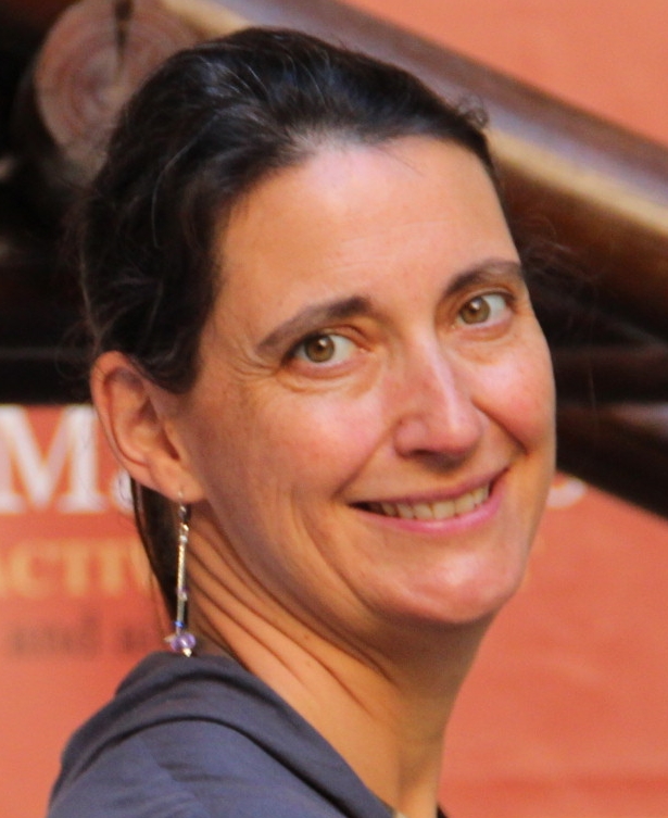 Portrait d'une femme de côté avec la tête tournée vers l'objectif de l'appareil photo. Elle a un sourire, des boucle d'oreille et elle a des cheveux foncé attaché en queue de cheval