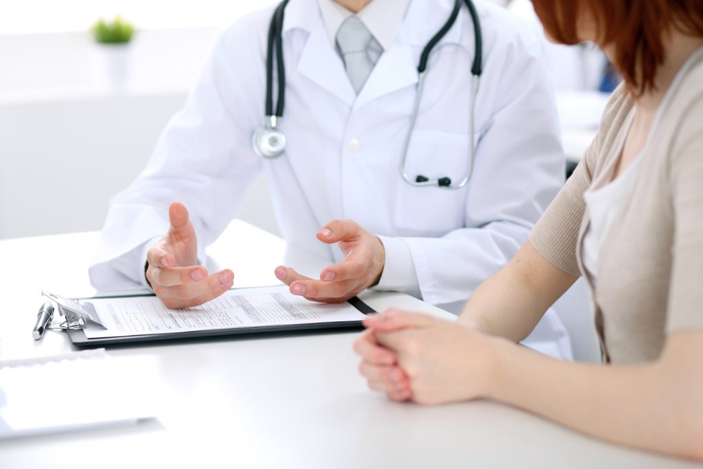 Consultation médicale entre un médecin en blouse blanche avec stéthoscope et une patiente, discussion autour d’un dossier médical sur un bureau blanc en cabinet de santé moderne