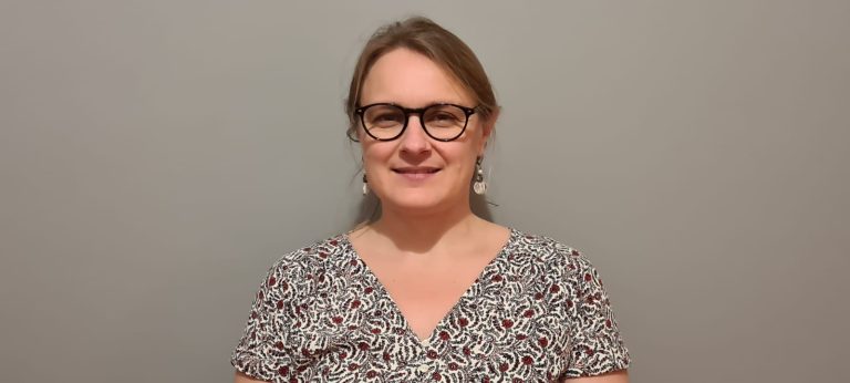 Portrait d’une femme souriante portant des lunettes et une robe imprimée, posant devant un mur gris uni