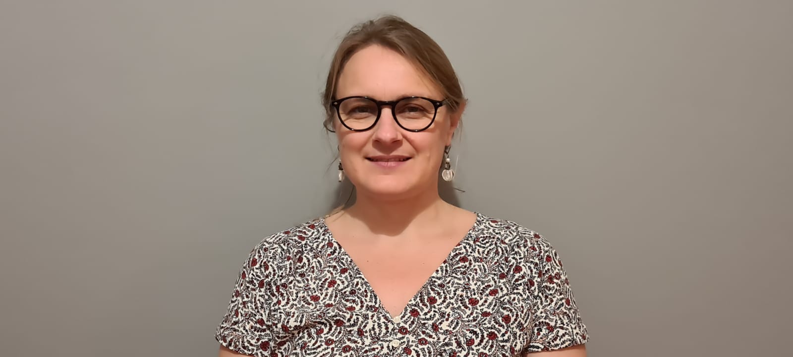 Portrait d’une femme souriante portant des lunettes et une robe imprimée, posant devant un mur gris uni