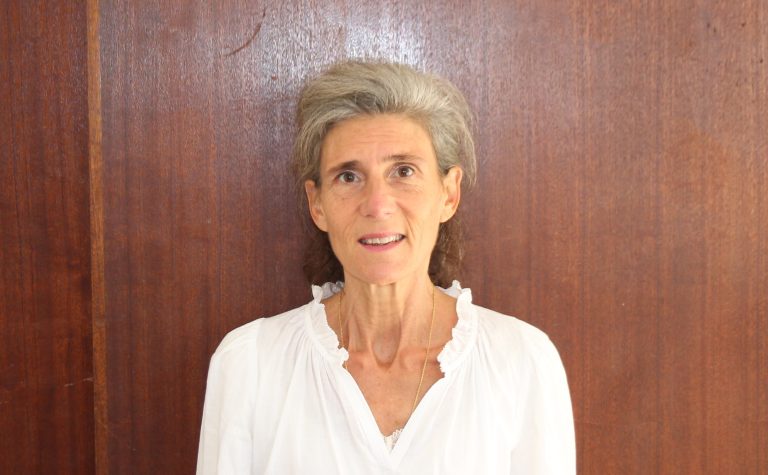 Femme souriante portant une chemise blanche, debout devant un fond en bois brun