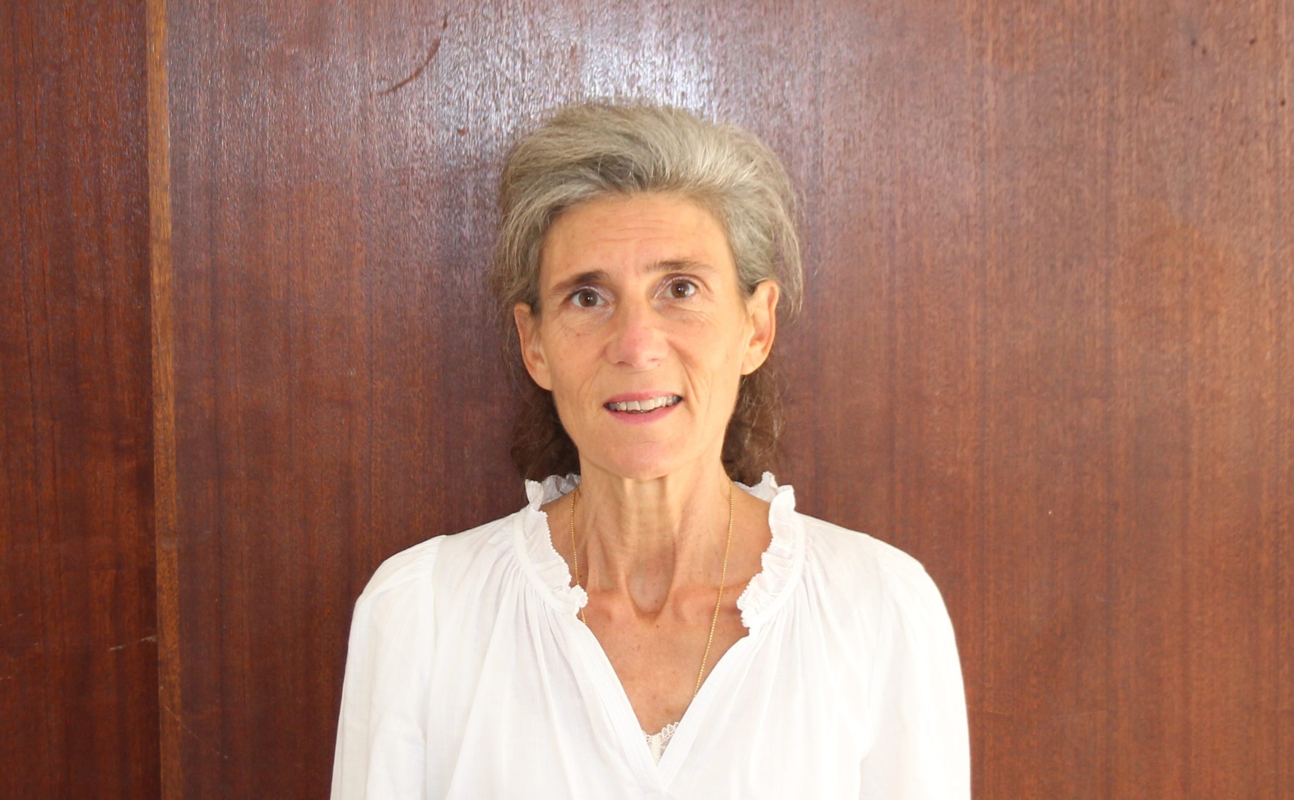 Femme souriante portant une chemise blanche, debout devant un fond en bois brun