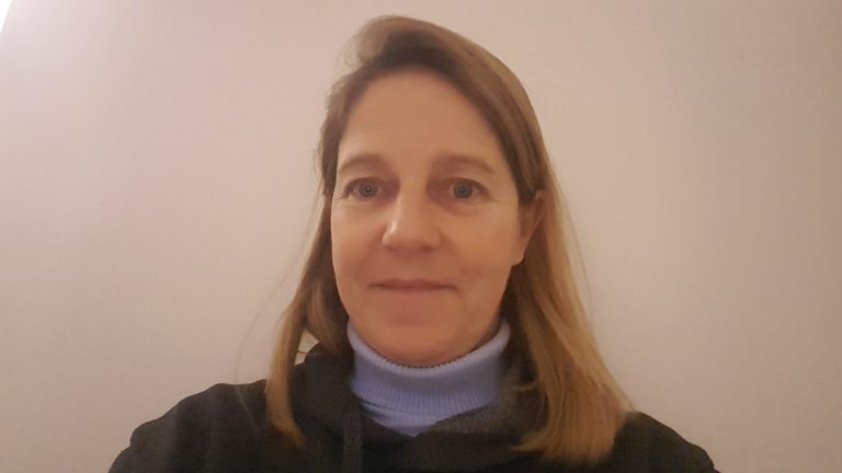 Portrait d’une femme aux cheveux blonds mi-longs, portant un pull foncé et un col roulé bleu clair, posant devant un mur uni dans une lumière douce