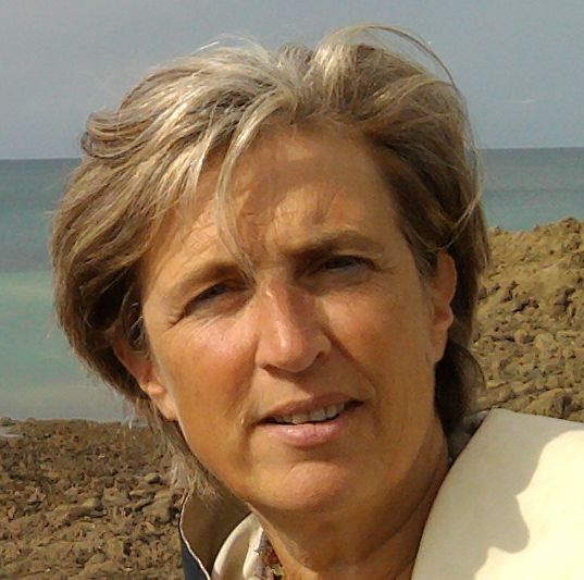 Portrait d'une femme aux cheveux blonds mi-longs, souriante, se tenant sur une plage ensoleillée avec la mer en arrière-plan