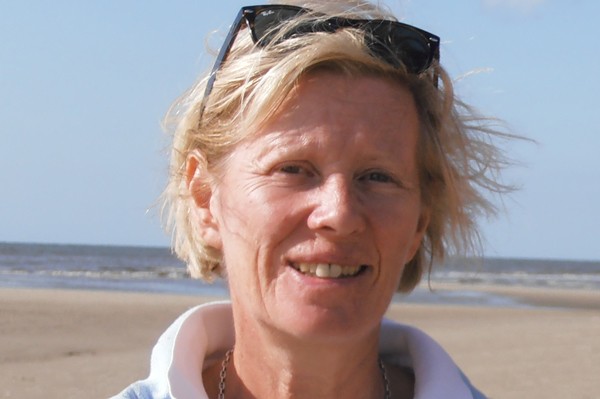 Portrait d'une femme souriante avec des cheveux blonds courts, portant des lunettes de soleil sur la tête, se tenant sur une plage ensoleillée avec la mer en arrière-plan.