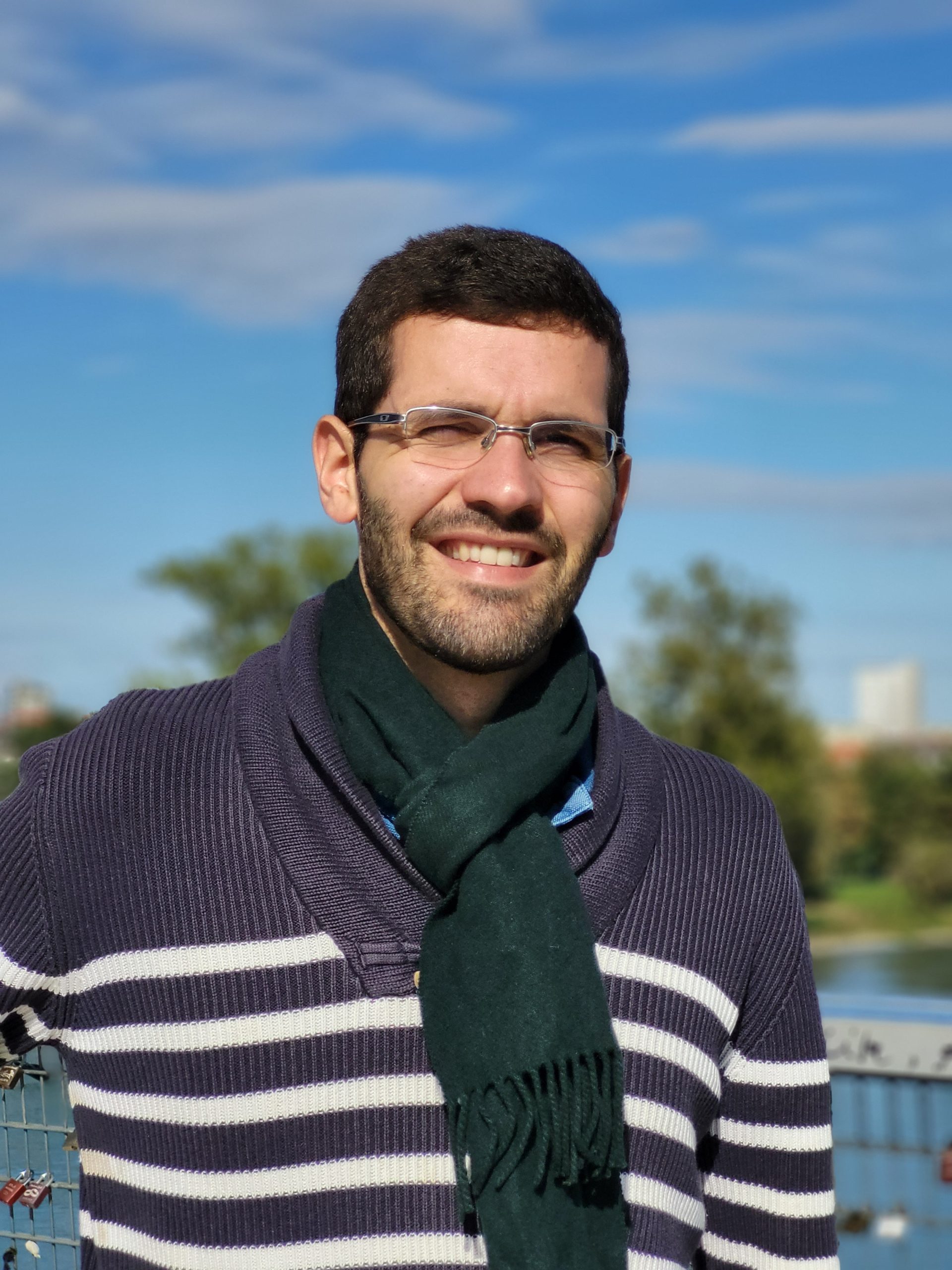 Homme souriant en pull rayé et écharpe verte, posant en extérieur par temps ensoleillé