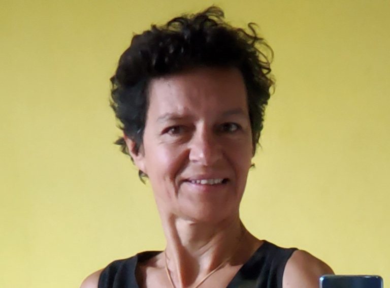 Portrait d’une femme souriante aux cheveux courts et bouclés, portant un débardeur noir, sur fond jaune uni, en lumière naturelle