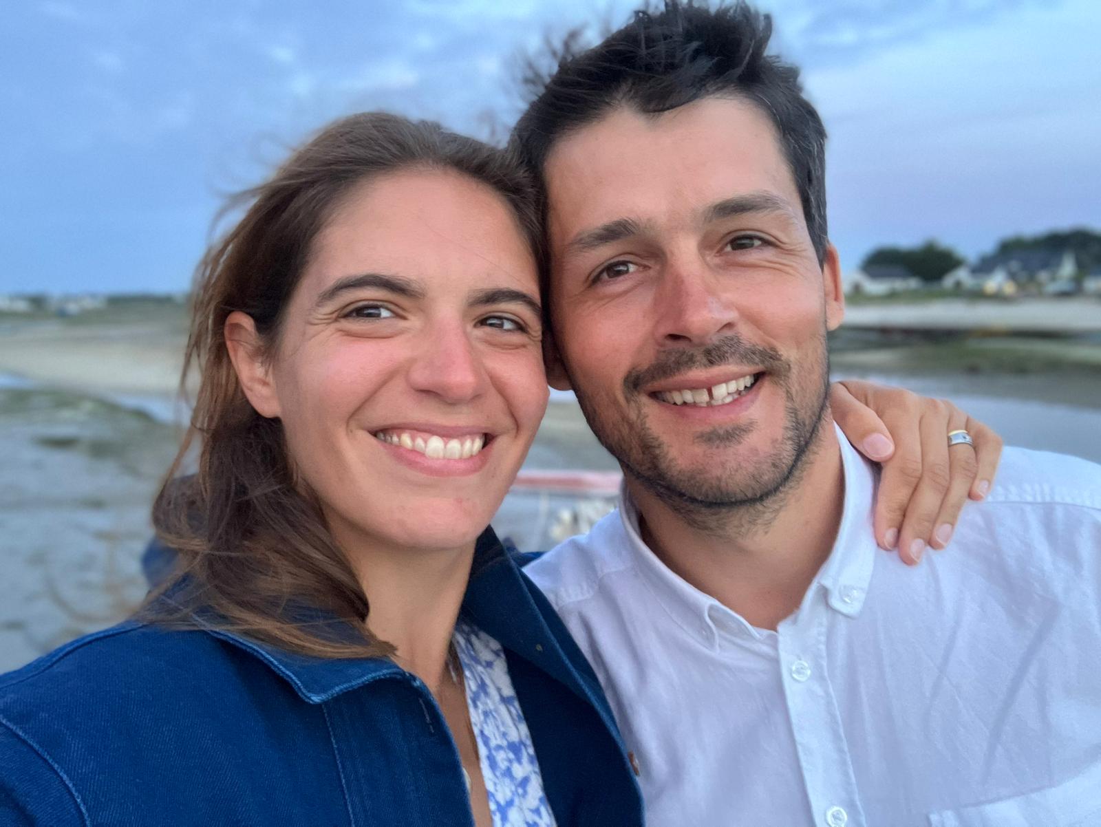 Couple souriant prenant un selfie au bord de la mer, bras autour des épaules, par une soirée d’été, avec un paysage côtier en arrière-plan