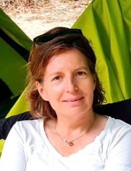 Delphine est une personne d'âge moyen et souriante. Elle est blanche de peau avec des cheveux cous de couleurs châtain claire avec un haut col en forme de V de couleur blanche