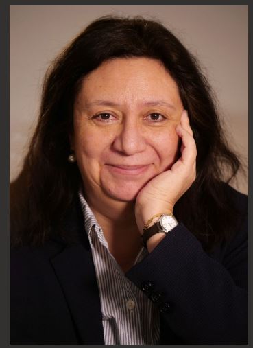 Portrait d'une femme souriante avec des cheveux longs et bruns, vêtue d'une veste bleue et d'une chemise à rayures, posant avec la main sur le menton.