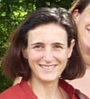 Portrait d'une femme souriante avec des cheveux bruns courts, portant un haut rouge, se tenant en extérieur avec un arrière-plan flou de verdure.