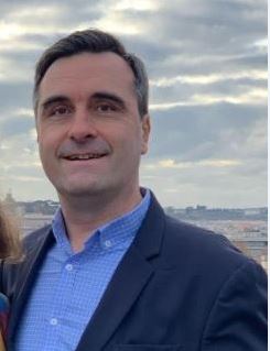 Portrait d'un homme souriant avec des cheveux bruns courts, portant une chemise bleue et une veste sombre, se tenant en extérieur avec un paysage urbain et un ciel nuageux en arrière-plan.
