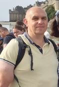 Portait d'un homme chave souriant avec un polo de couleur jaune claire et une sac a dos gris et noir. En arrière plan nous pouvons observer quelques personnes se baladant