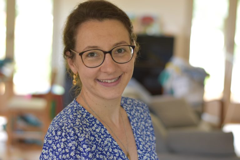 Portrait d’une femme souriante portant des lunettes, une robe bleue à fleurs blanches et des boucles d’oreilles dorées, posant dans un intérieur lumineux avec canapé en arrière-plan