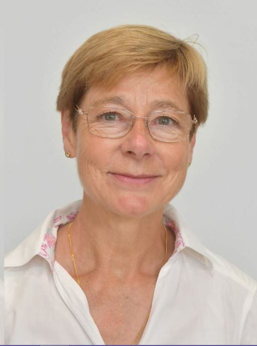 Portrait d’une femme souriante aux cheveux blonds courts, portant des lunettes, une chemise blanche et un collier discret, sur fond clair