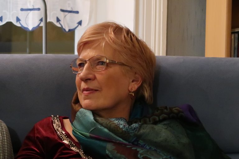 Portrait d’une femme élégante portant des lunettes et un châle coloré, assise sur un canapé bleu, regardant vers la gauche avec un sourire paisible