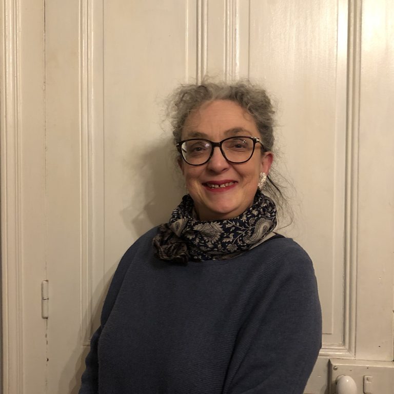 Portrait d’une femme souriante portant des lunettes, une écharpe à motifs floraux et un pull bleu, posant devant une porte en bois clair