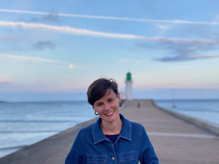 Jeune femme souriante en veste en jean, posant sur une jetée face à la mer avec un phare en arrière-plan au coucher du soleil