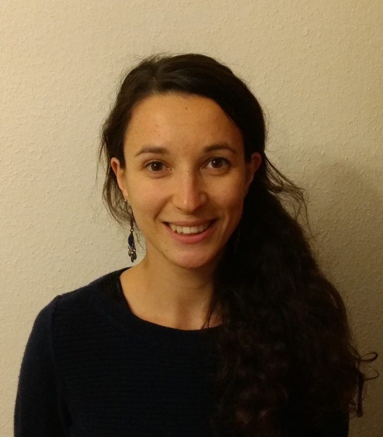 Portrait d'une femme souriante aux cheveux longs et bouclés, attachés sur le côté, portant un pull foncé et des boucles d'oreilles pendantes, devant un mur clair
