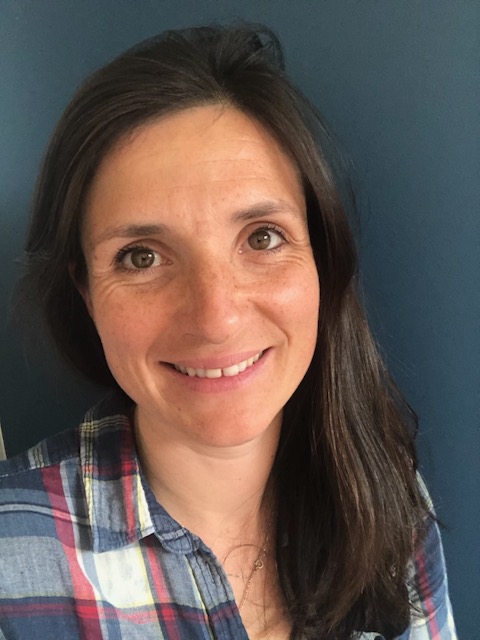Portrait d'une femme souriante avec des cheveux bruns mi-longs, portant une chemise à carreaux bleus et rouges, se tenant devant un fond bleu uni.