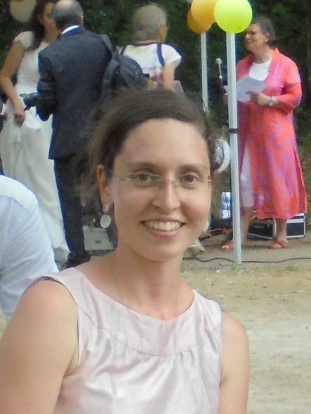 Portrait d'une femme souriante avec des cheveux bruns et des lunettes, portant une robe sans manches de couleur claire, se tenant en extérieur lors d'un événement festif avec des ballons jaunes et des personnes en arrière-plan.