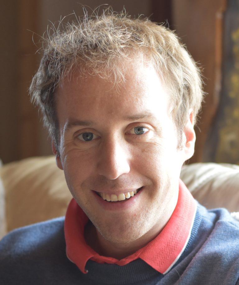 Homme souriant assis sur un canapé, en pull bleu et polo rouge, regardant l’objectif dans une lumière douce d’intérieur