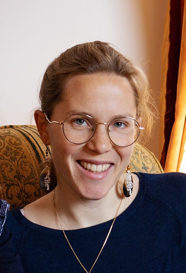 Portrait d’une femme souriante aux lunettes rondes, portant un pull bleu marine et de longues boucles d’oreilles, assise dans un fauteuil à motif classique, en intérieur