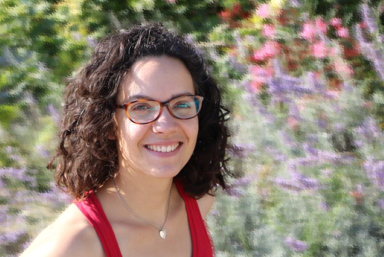 Femme souriante aux cheveux bouclés et lunettes, portant un débardeur rouge, posant devant un jardin coloré en arrière-plan flouté