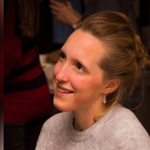 Portrait d'une femme souriante avec des cheveux blonds attachés en chignon, portant un pull gris et des boucles d'oreilles, regardant sur le côté avec une expression joyeuse.